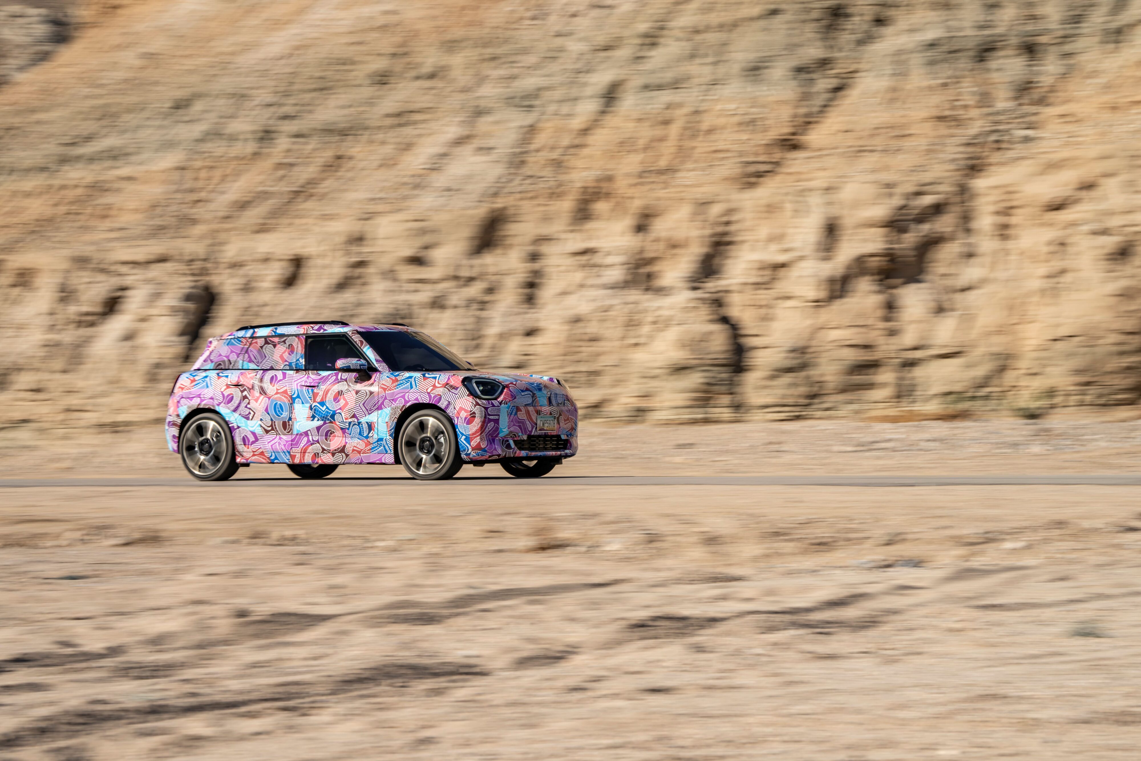 Front three quarters view of a camouflaged prototype MINI Aceman EV in a desert surrounding.