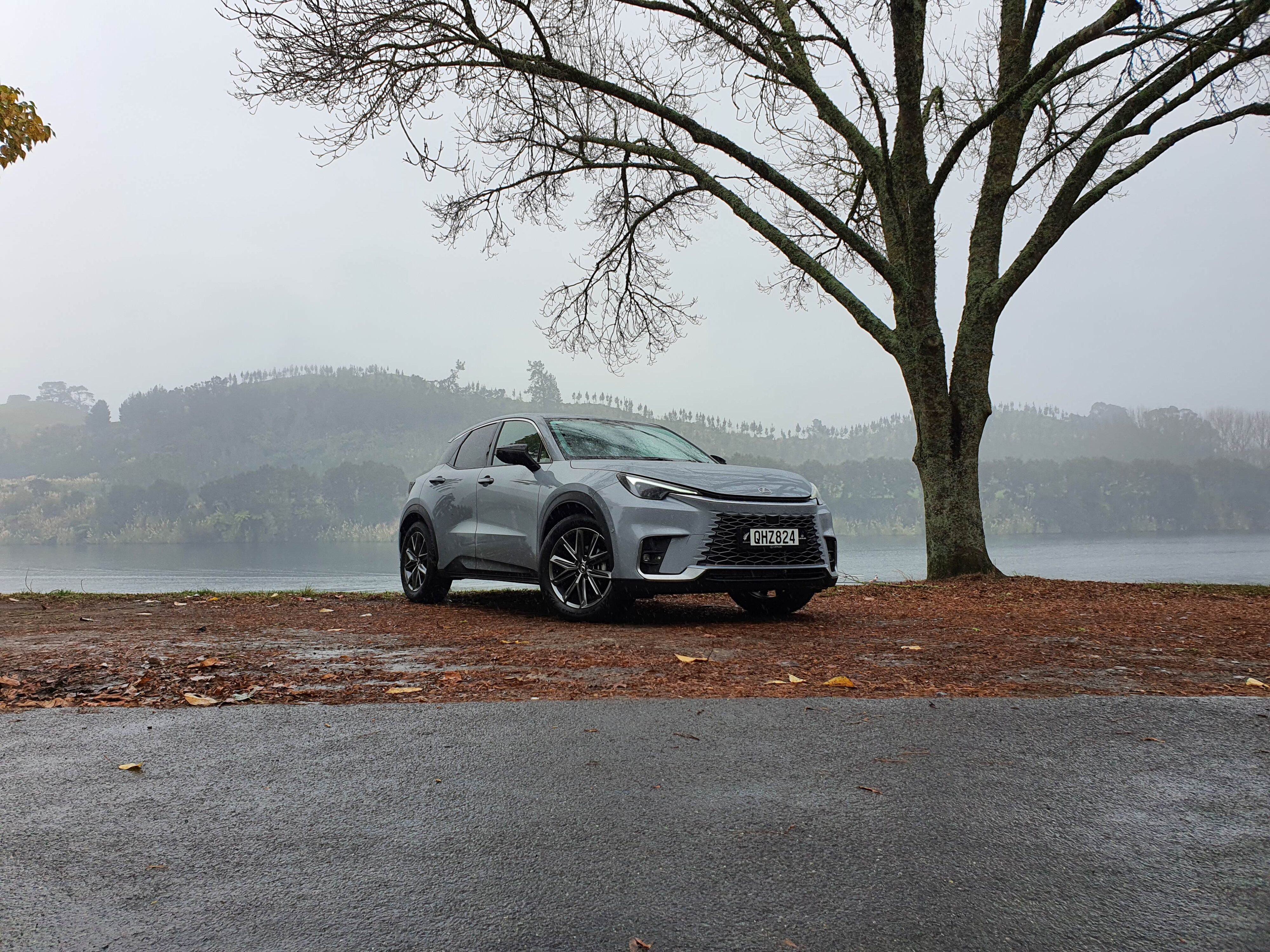 Front three quarters view of a Lexus LBX Relax AWD in Concrete Grey with a misty background of bare trees and Lake Karapiro.