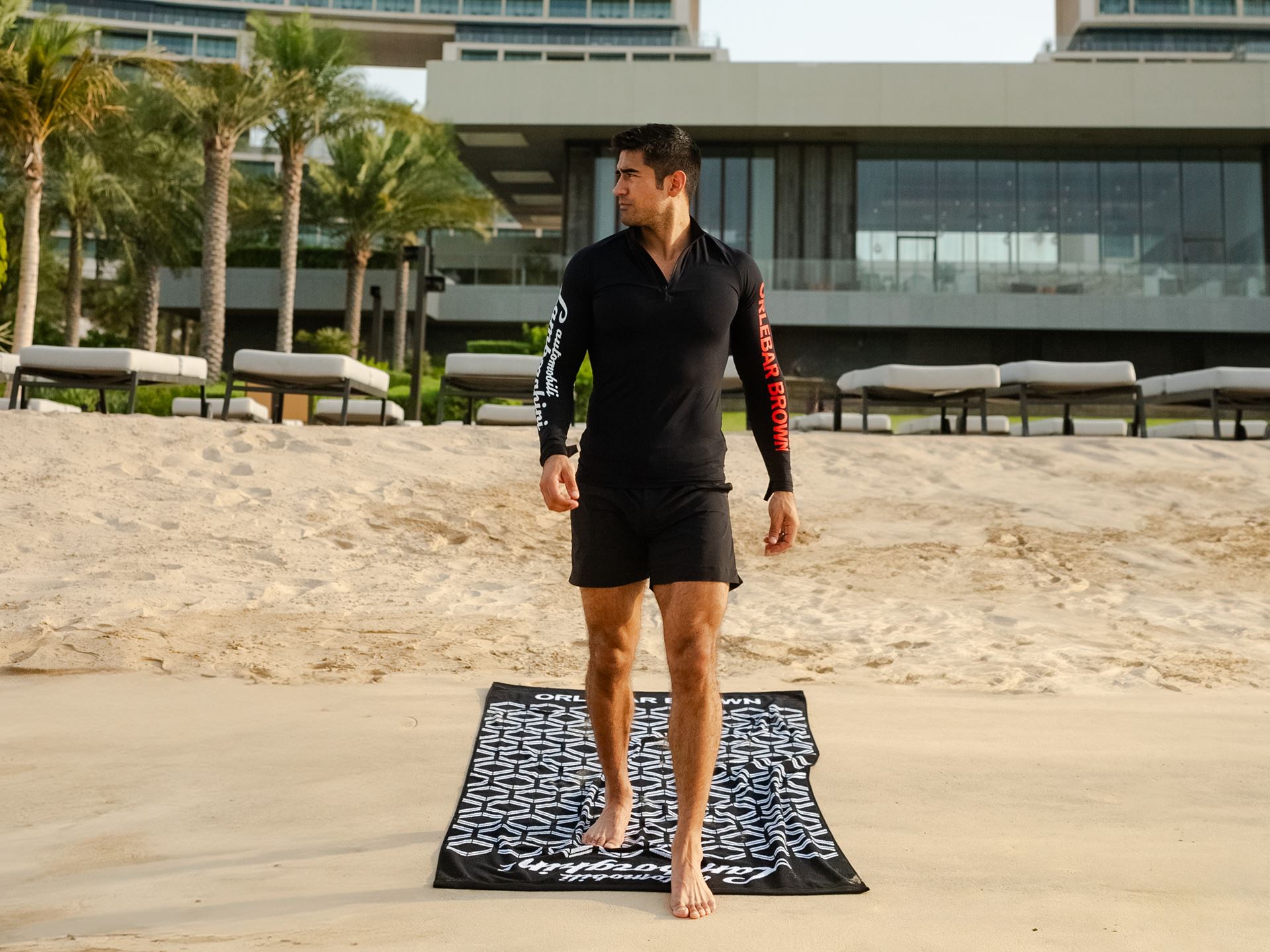 A man dressed in the Lamborghini by Orlebar Brown collection walks off a Lamborghini x Orlebar Brown beach towel.
