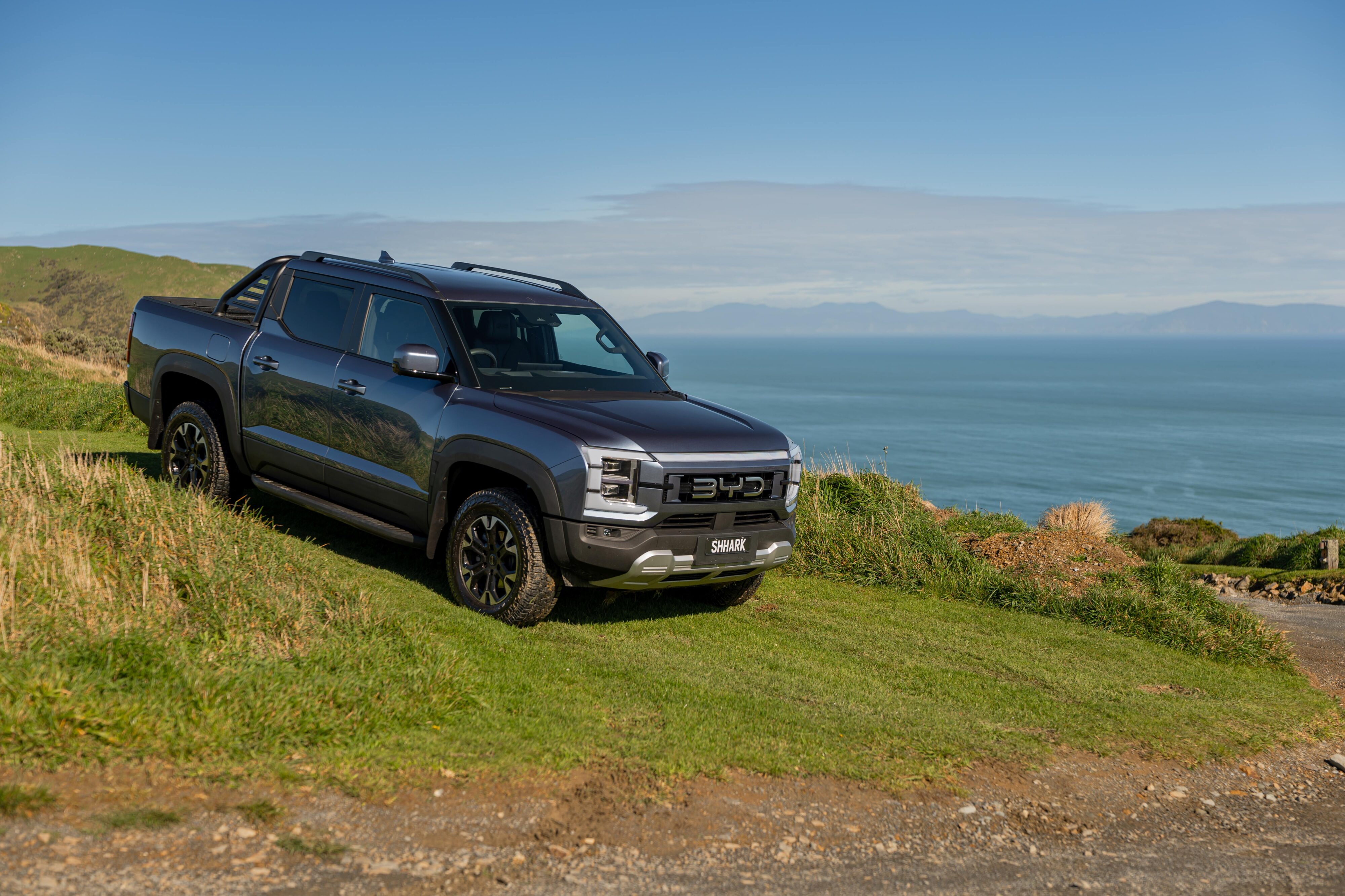 A front three quarters view of a BYD Shark 6 on a hill with the ocean in the background.