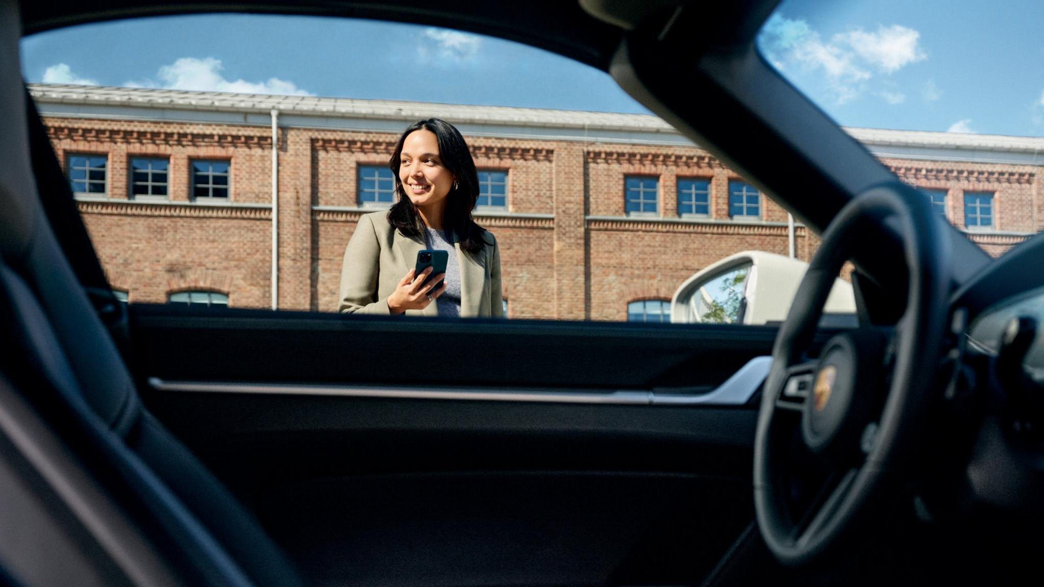 A woman holding a phone as seen through the window of a Porsche car.