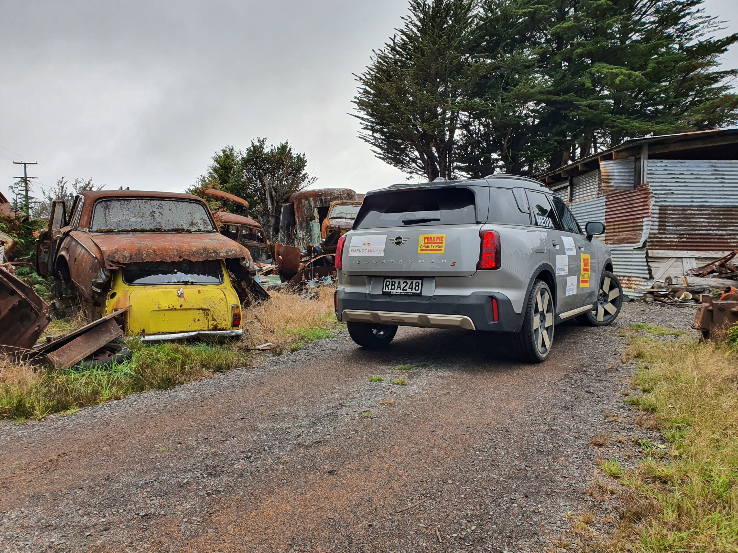 Rear three quarters view of a 2025 Mini Countryman S with Employee Lab and Mortgage Lab branding on the side parked in Smash Palace, Horopito with a classic yellow Mini visible on the left.