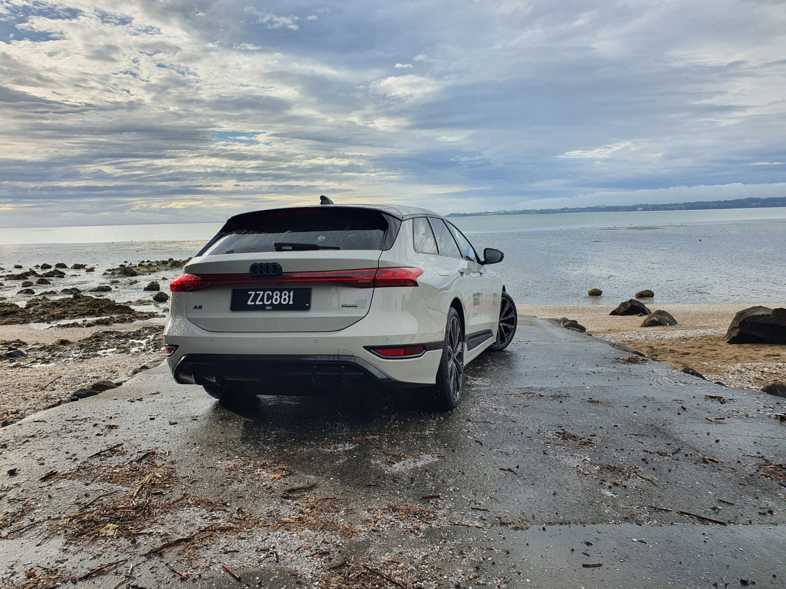 Rear three quarters view of an Audi A6 Avant e-tron S-Line quattro in Siam Beige with the ocean in the background.