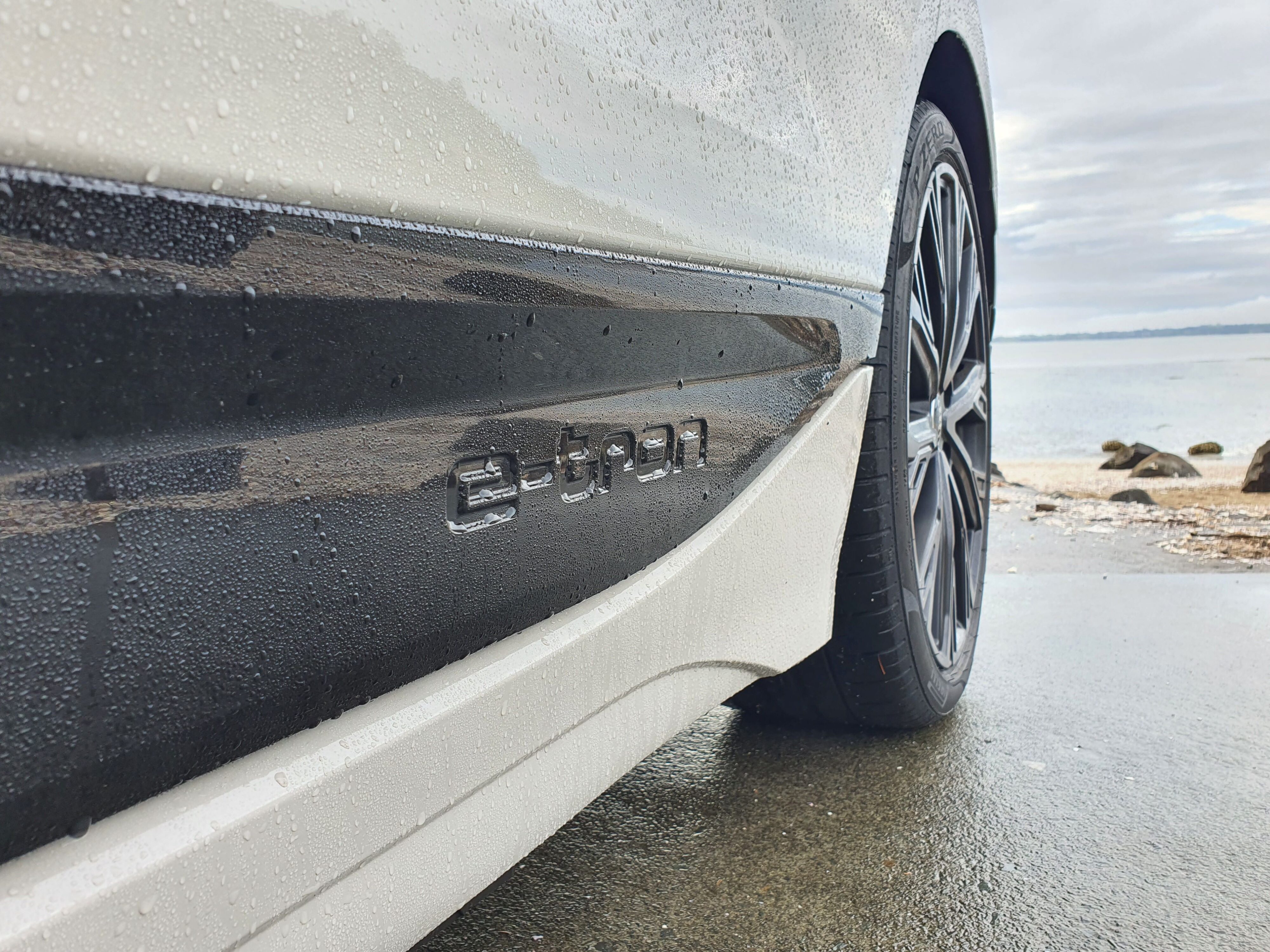 e-tron embossed badging on the left rear side of an Audi A6 Avant e-tron S-Line quattro in Siam Beige with the ocean in the background.