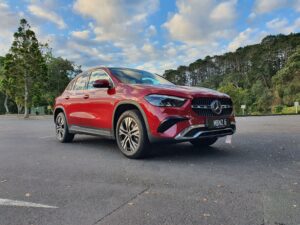 Front three quarters view of a 2025 Mercedes-Benz GLA250e in Patagonia Red Metallic with trees in the background.