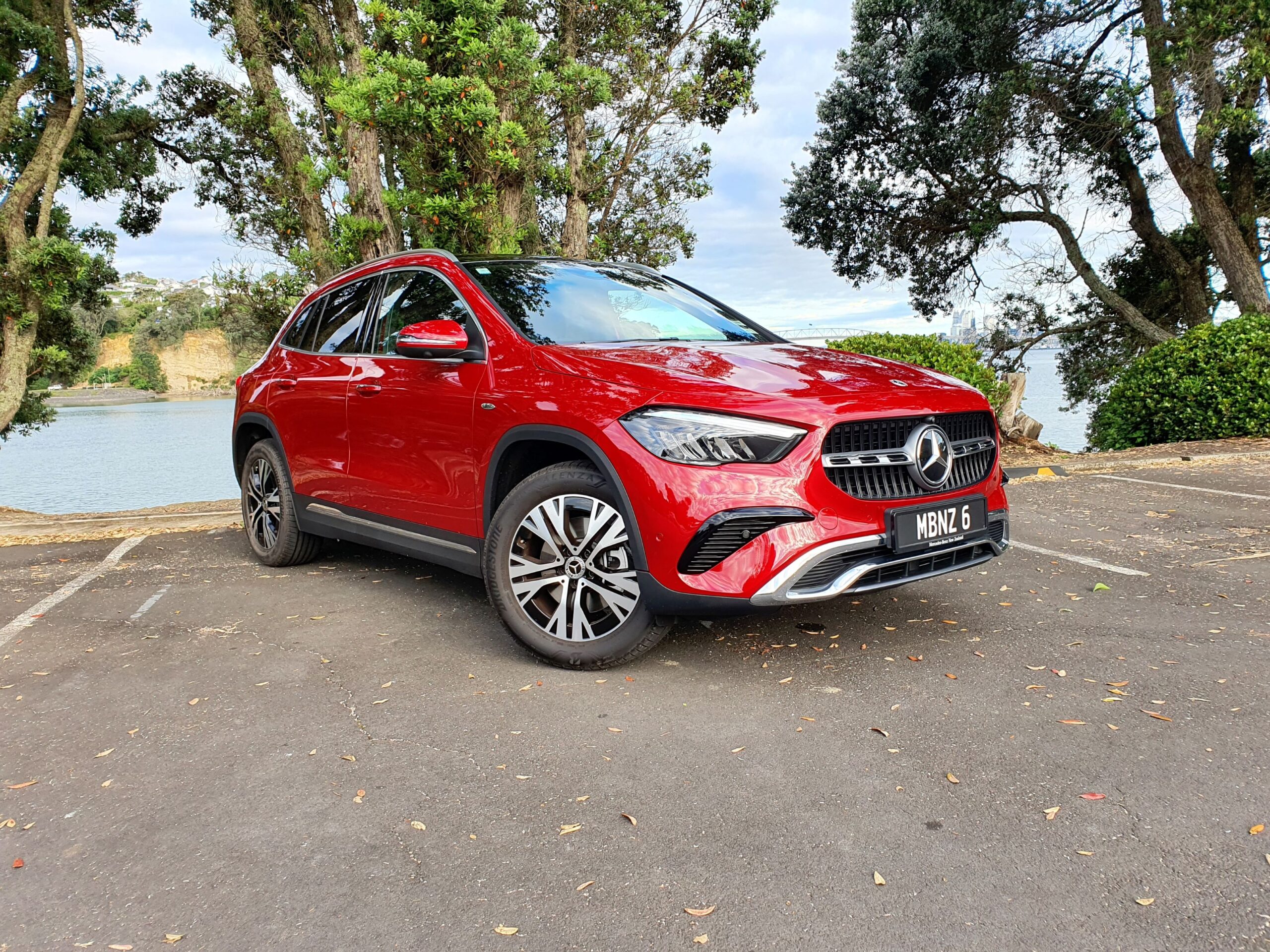 Front three quarters view of a 2025 Mercedes-Benz GLA250e in Patagonia Red Metallic with the ocean in the background.
