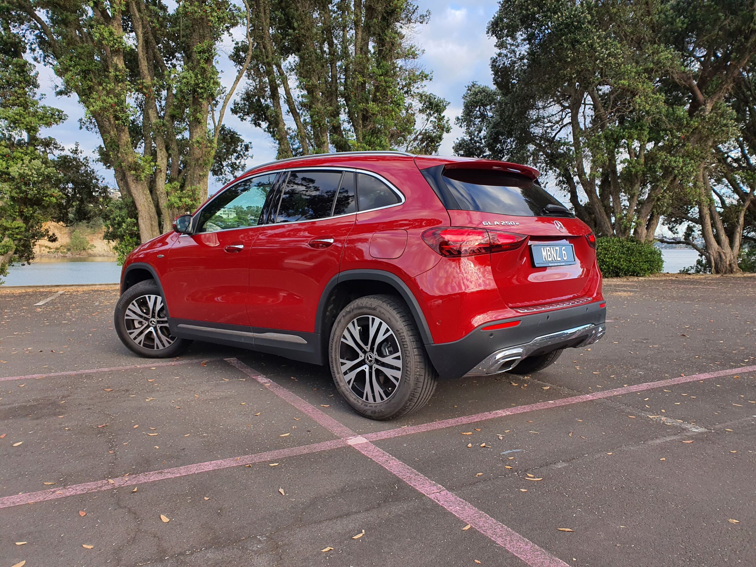 Rear three quarters view of a 2025 Mercedes-Benz GLA250e in Patagonia Red Metallic with the ocean in the background.