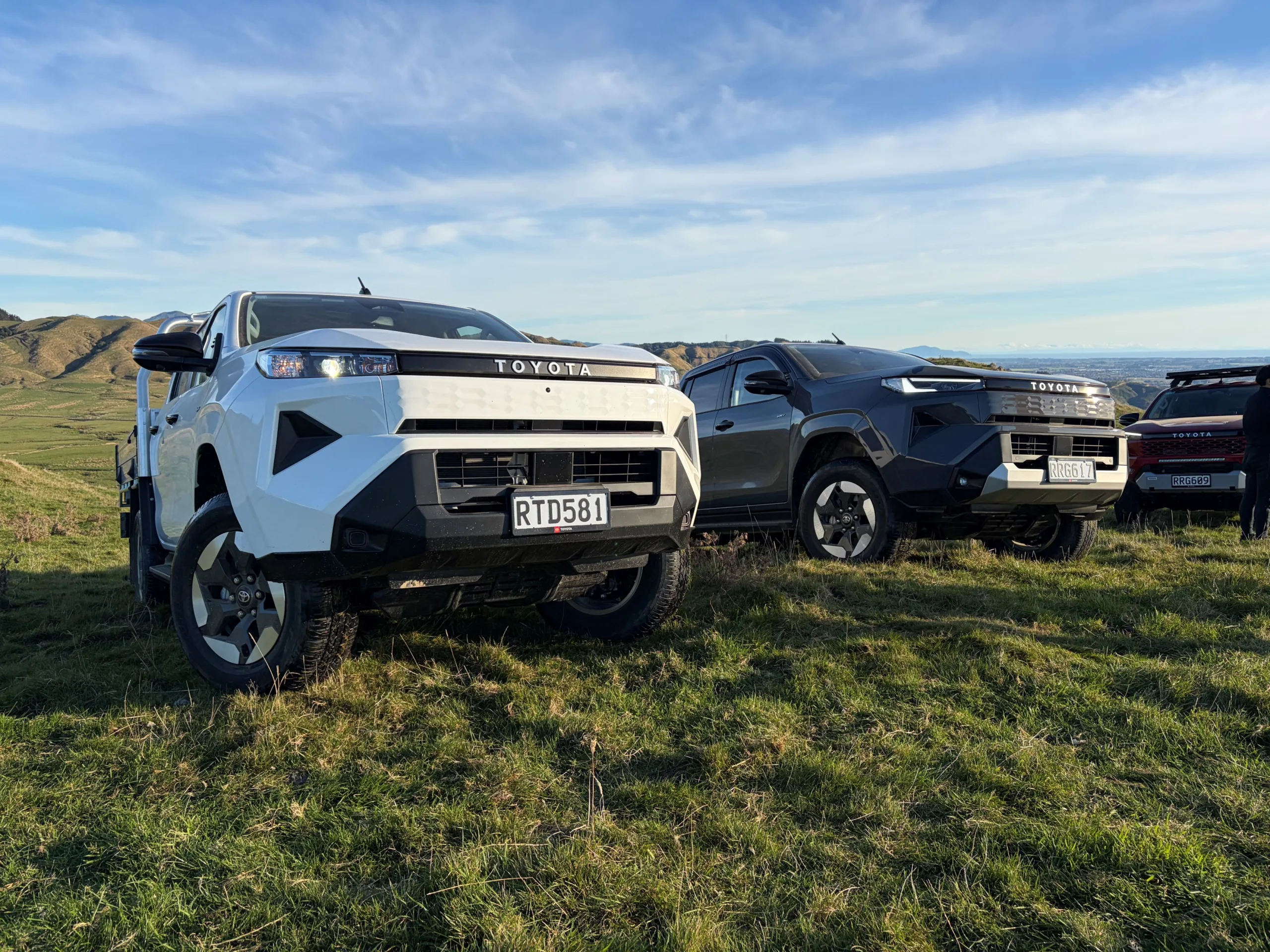 Close up front three quarters view of a 'Glacier White' 2026 Toyota Hilux SR BEV with an 'Ash' coloured 2026 Toyota Hilux SR5 BEV in the background. Taken on the launch of the ninth-generation Hilux in the Manawatu, New Zealand.