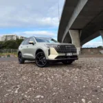 Front three quarters view of a 2026 GWM Haval H6 HEV in Astral Pearl, photographed against an urban backdrop.