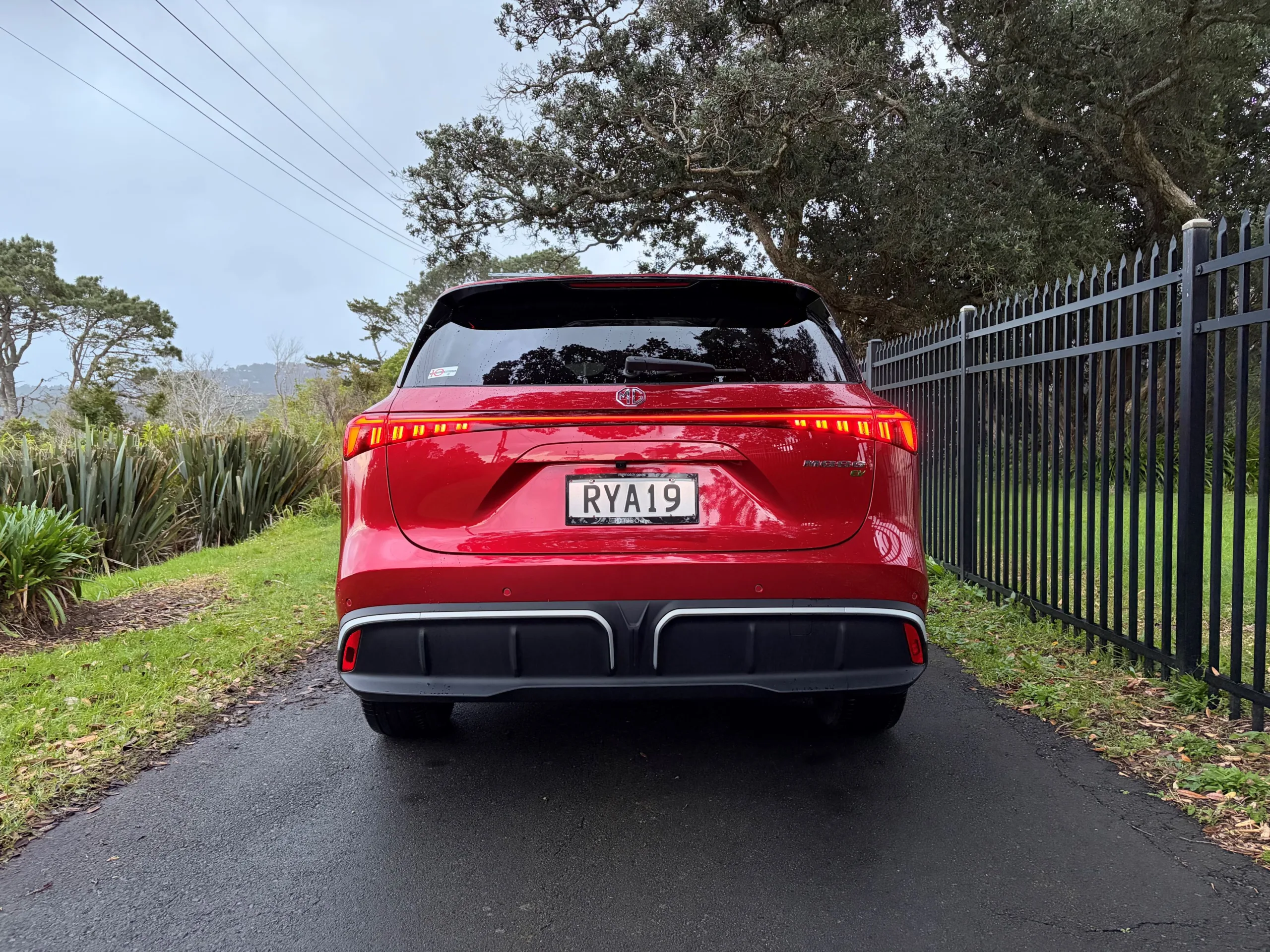 Rear straight on view of a 2026 MG S6 EV AWD in Diamond Red, nature/greenery in the background.